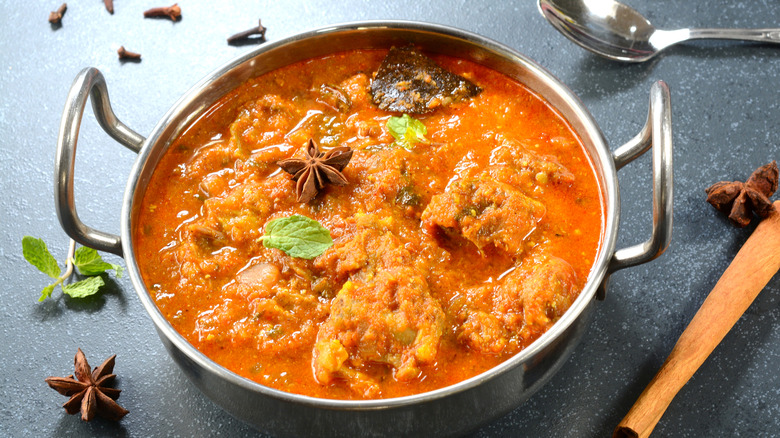 An Indian curry in a steel bowl surrounded by a scatter of whole spices