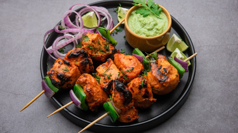 A black plate with chicken kebabs on skewers served with a small bowl of cilantro sauce and raw onion rings and lime wedges