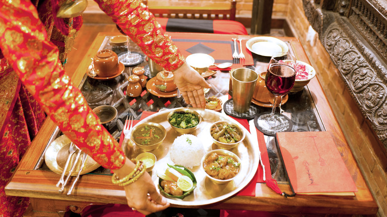 A traditional table set with Indian food, pickles, drinks and the hands of a server dressed in traditional finery
