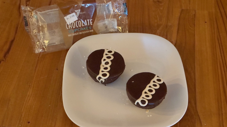 Two chocolate cupcakes with a decorative white icing swirl on a square white plate next to a 7-Select wrapper