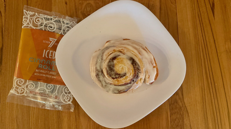 An iced cinnamon roll on a square white plate next to a plastic 7-Select wrapper