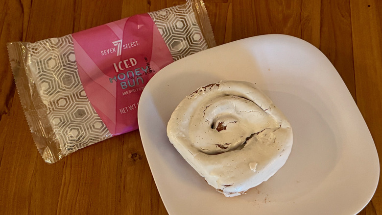 A heavily iced honey bun on a square white plate next to a plastic wrapper