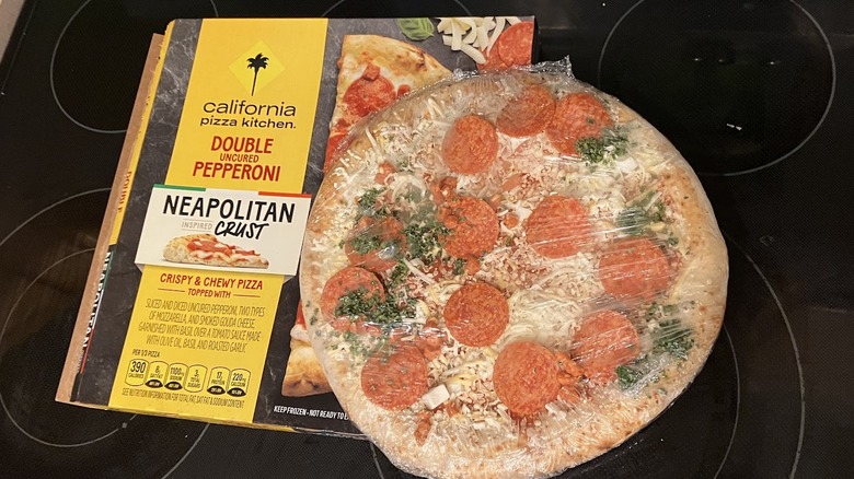 Frozen pepperoni pizza with green herbs sits atop a box of California Pizza Kitchen brand pizza.