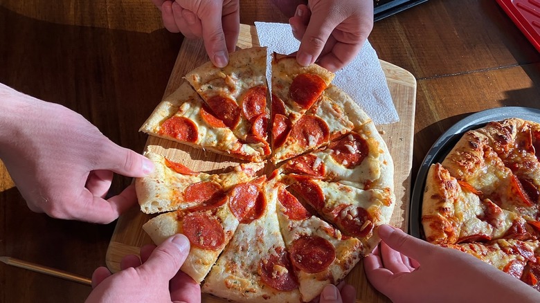 Several hands reach for a slice of a round pepperoni pizza. Another sliced pizza waits beside it.