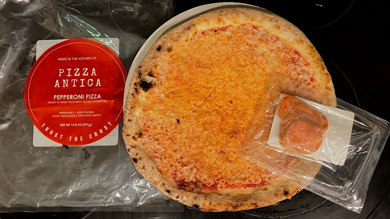 A large, round orange frozen pizza with a separate packages of pepperoni slices, next to the pizza's plastic packaging.