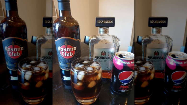 A Cuba libre cocktail sits on a table in glass with ice, with a Pepsi can to the right. Bottles of liquor are placed on the table behind it.