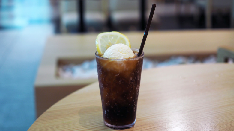 A glass of Pepsi with a scoop of vanilla ice cream sits on a wooden table. A lemon wedge and straw are sticking out of the glass.