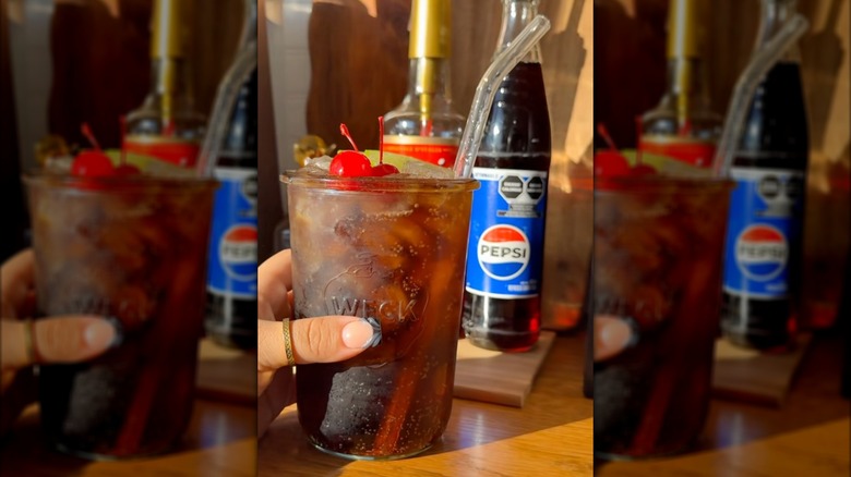 A closeup of a Shirley temple created from Pepsi, Garnishes like cherry and lime rest on the top, with glass Pepsi bottle in background.