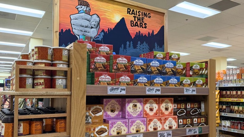 Snack bar end cap display at Trader Joe's under hand-painted sign that reads "Raising the Bars" with image of mountains at sunset
