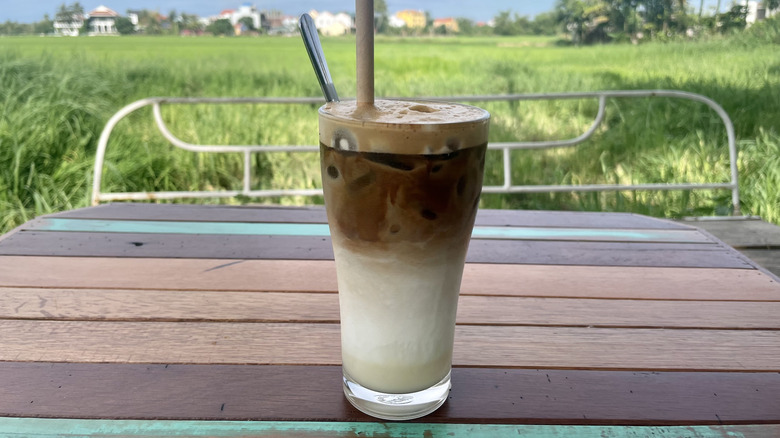 Bac Xiu in clear glass with straw sits on wooden table overlooking rice fields