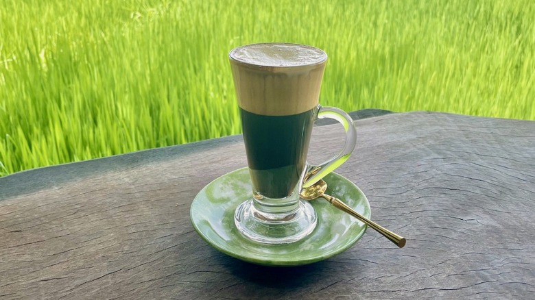 Hot egg coffee with golden spoon sits on old wooden table overlooking rice field