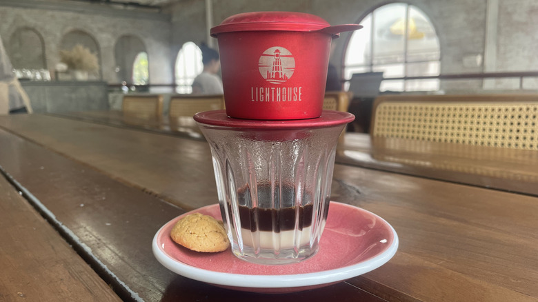 Red Lighthouse phin filter sitting atop clear glass with condensed milk and coffee by cookie