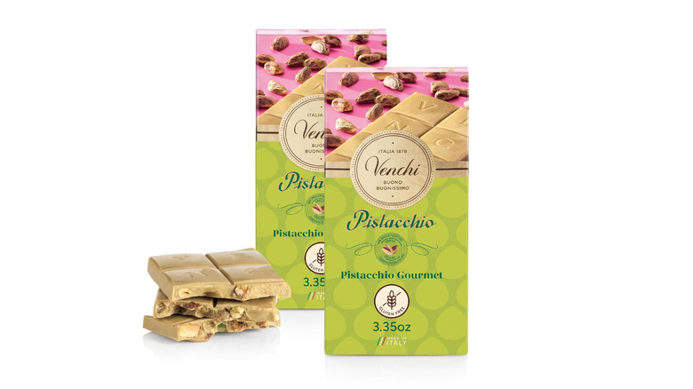 Two Venchi Pistachio Gourmet White Chocolate Bars in package alongside stack of pistachio white chocolate squares on whitebackground
