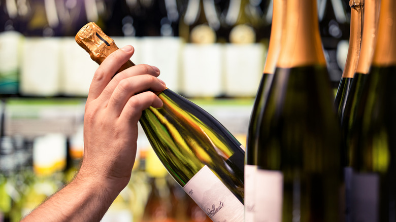 Man's hand grabs a bottle of sparkling wine from row of bottles on shelf in alcohol store.