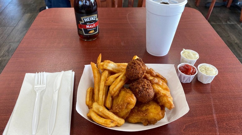 Classic fish and chips in a paper basket on a table, next to a napkin, plasticware, and cups of sauces.