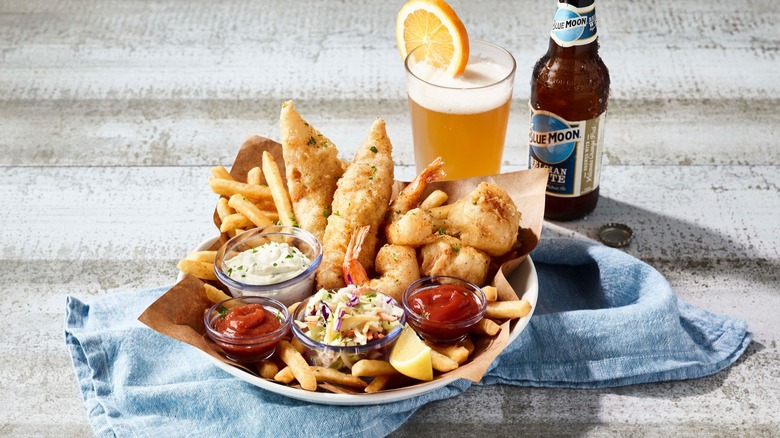 Bonefish Grill's Crispy Cod on a platter with fries, cole slaw, tartar and cocktail sauce, next to a glass and bottle of Blue Moon beer.
