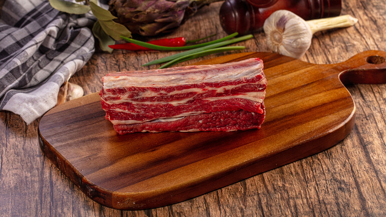 Fresh raw beef short ribs on rustic wooden cutting board surrounded by garlic, red chili peppers, green onions, bay leaves, artichoke, pepper mill