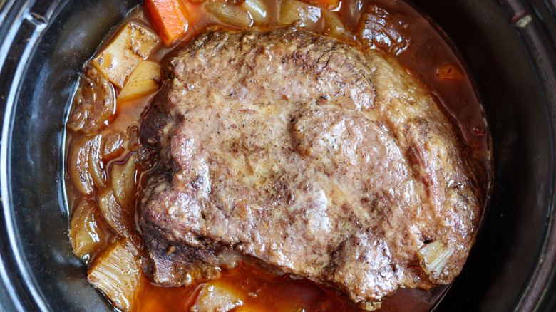 A homemade pot roast cooking inside of a crockpot with onions and carrots