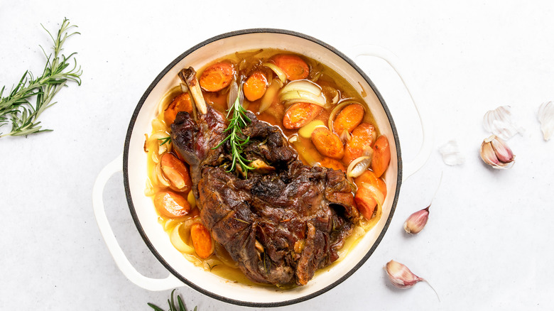 Lamb shoulder with onions and carrots in a cast iron pan surrounded by garlic cloves and rosemary sprigs