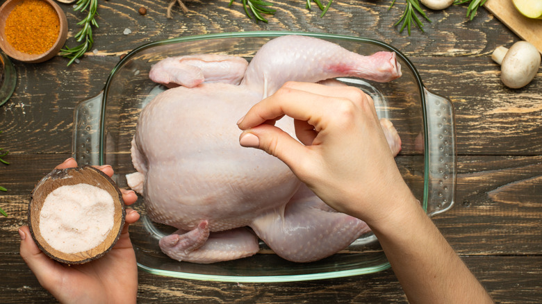 Top view of hands sprinkling whole raw chicken in glass dish with salt