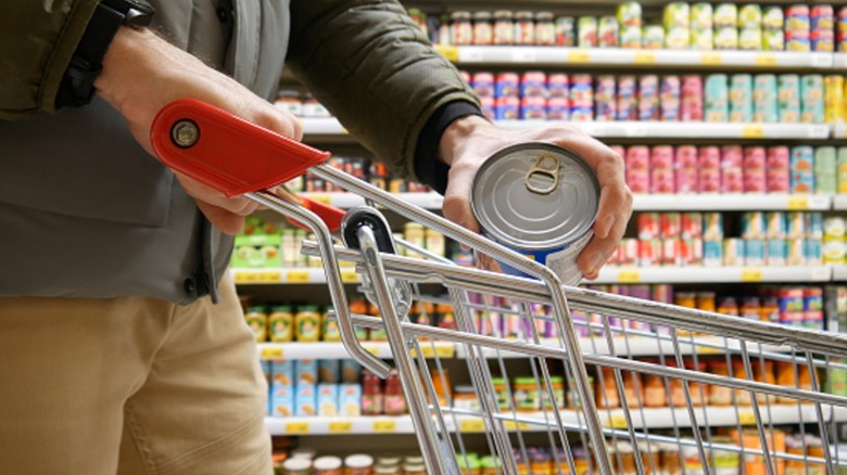 Close-up of a shopping cart in canned food aisle and a male shopper shown putting a can into it