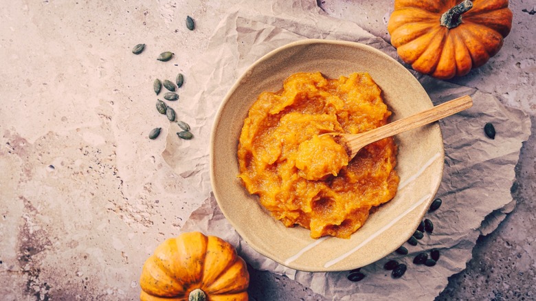 orange pumpkin puree displayed in a bowl with a spoon on a table, with mini pumpkins and a few scattered seeds beside it, aerial view