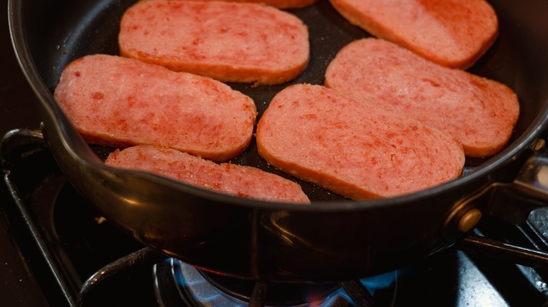Slices of Spam frying in a black pan over a gas stove with a blue flame