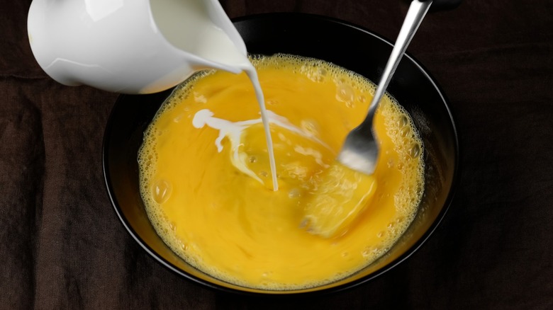 Hand seen pouring heavy whipping cream while mixing eggs with fork