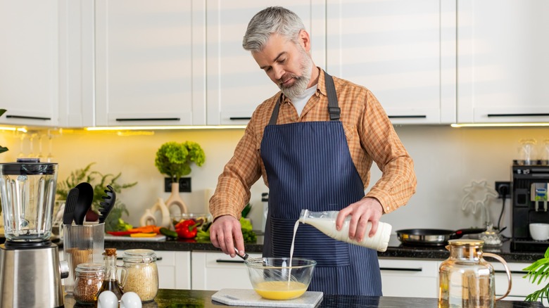 Middle-aged man adds milk to beaten eggs and whisks bowl in kitchen
