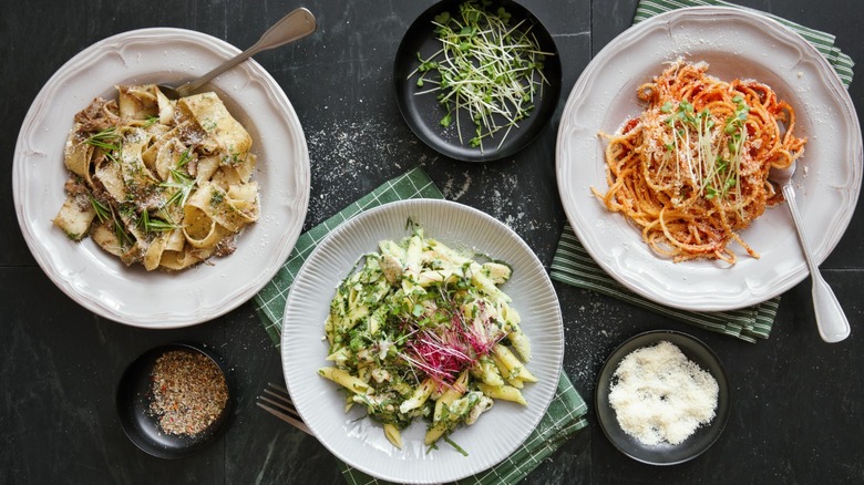 three different bowls of pasta with meat, greens, and red sauce alongside small dishes of garnish and parmesan cheese.