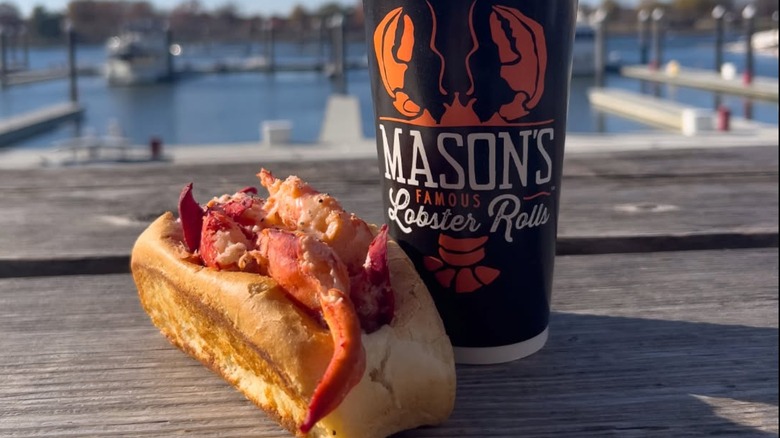 Lobster roll beside a drink with the store logo on it sitting on a wooden dock with the sea in the background at Mason's Famous Lobster Rolls