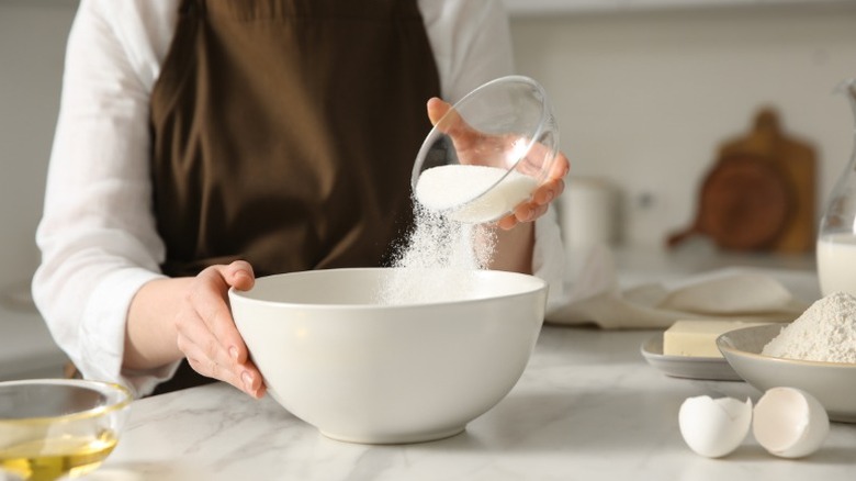 a person pours a small glass bowl of sugar into a large mixing bowl