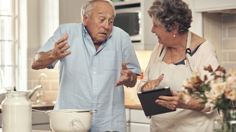 an elderly man and woman look confused while looking at a recipe in the kitchen