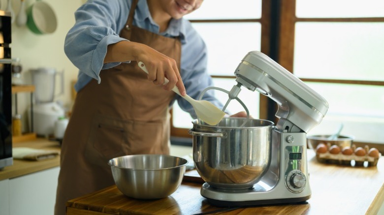 a man in an apron uses a spatula to scrape the bowl of a stand mixer in a kitchen