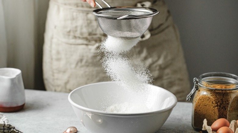 a person in an apron sifts flour into a mixing bowl on a counter next to a jar of brown sugar and carton of eggs