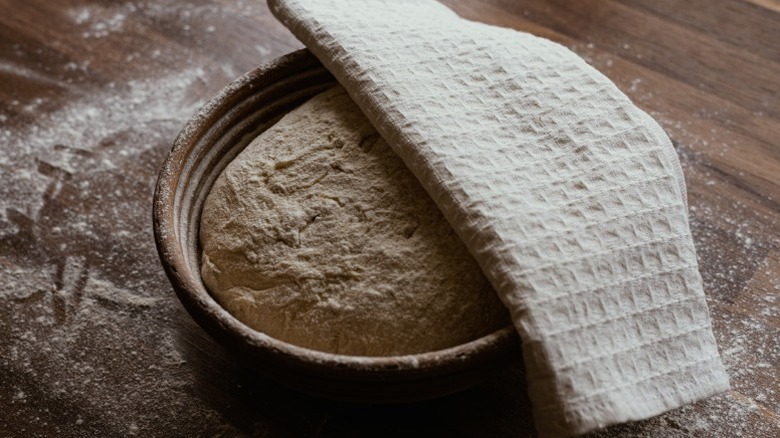 a bowl of dough half-covered with towel sits on a floured wooden surface