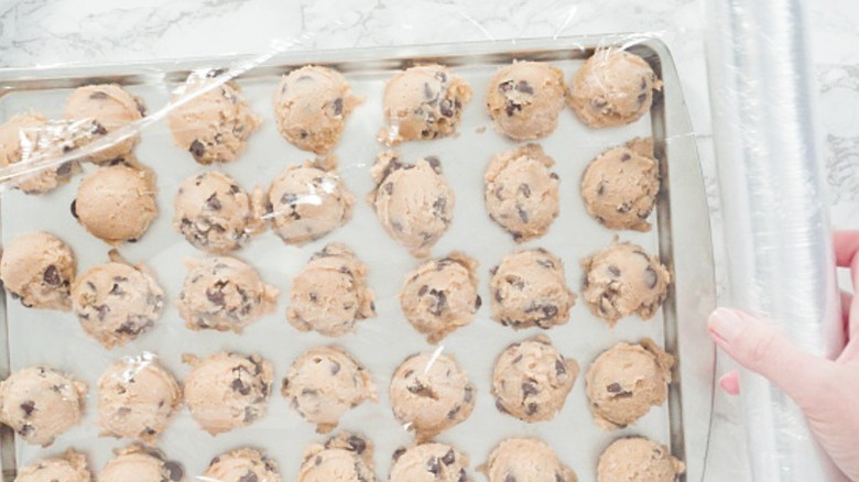 a hand pulls plastic wrap over a baking tray full of chocolate chip cookie dough scoops