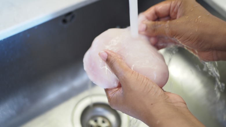 Hands washing raw chicken breast in a kitchen sink