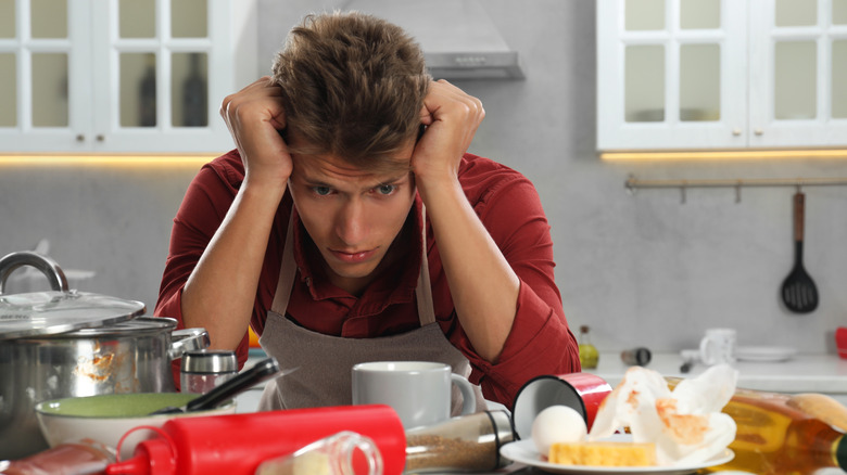 Upset man in messy kitchen. Many dirty dishware, utensils and food leftovers on table