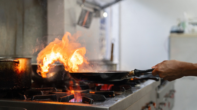 A chef flames a pan dramatically in a commercial kitchen, creating a high-heat fire while cooking steak.