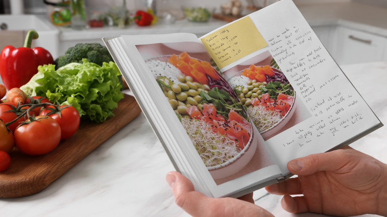 Closeup of man;s hands with recipe book at white marble table in kitchen, in front of wooden board with fresh produce