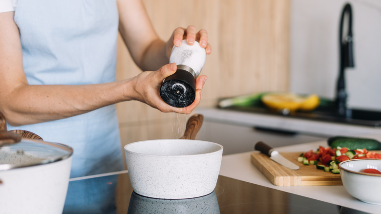 A woman adds black pepper to a white pot on the stovetop while preparing a vegetable dish in her modern kitchen.