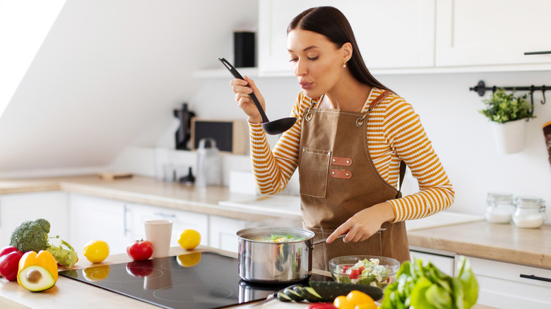 Woman blowing on black ladle of soup before tasting it while cooking alone in bright, white kitchen