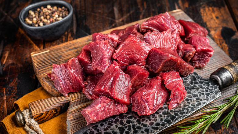 Chunks of lean red venison meat on wooden cutting board with knife and ramekin of black pepper