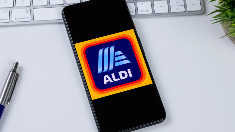 A smartphone showing the Aldi logo with a keyboard, plant and, pen in the background
