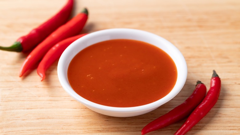 Sriracha sauce in a white ramekin on a wooden background accompanied by fresh chili peppers