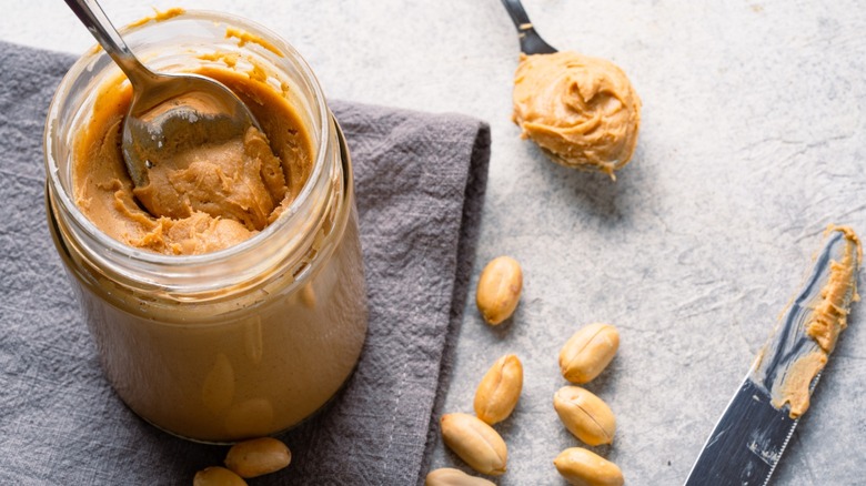 Open jar of creamy peanut butter sitting on a grey towel with a spoon in it, accompanied by whole peanuts, another spoon of peanut butter, and a knife covered in peanut butter