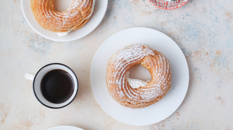 An overhead shot of a puff pastry donut filled with cream, on a white plate, next to a cup of coffee.