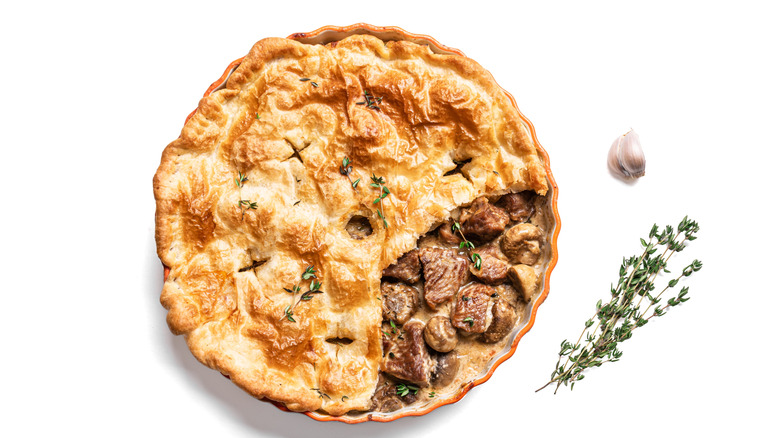 A meat and mushroom pot pie topped with puff pastry, a slice removed to show the filling, isolated on a white background.