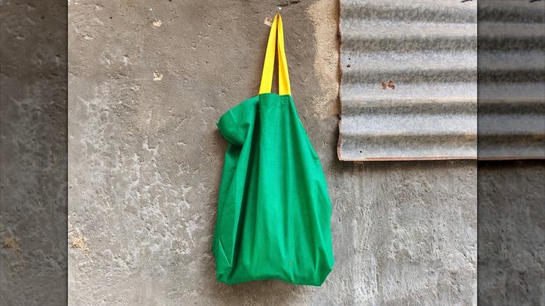 Green reusable cloth bag with yellow handles hanging on a textured cement wall